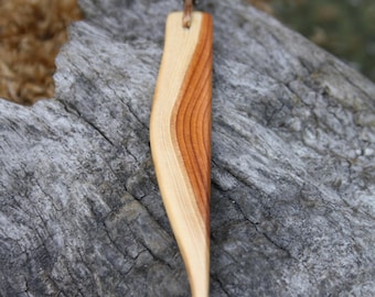 May include: A handcrafted wooden pendant necklace. The pendant is a curved, elongated shape with a light and dark wood grain pattern. It hangs from a brown leather cord with a silver-colored metal accent. The necklace is displayed on a weathered wooden surface.