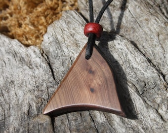 May include: A wooden pendant necklace with a triangular shape. The pendant is a warm brown color and is suspended from a black cord. A small red bead accents the cord. The necklace is displayed on a piece of weathered wood.
