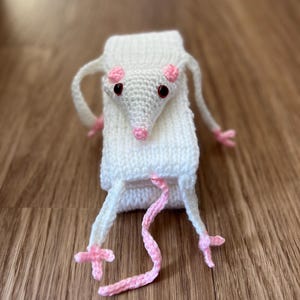 May include: A handmade white knitted rat with pink accents. The rat has a long, pink tail and pink paws. The eyes are black, and the ears and nose are pink. The rat is made of soft yarn and is likely a toy or decorative item.