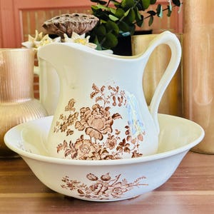 May include: A cream-colored ceramic pitcher and matching basin set featuring a detailed brown floral transferware pattern. The pitcher has a curved handle and a pouring spout, while the basin is wide and shallow, both decorated with flowers.