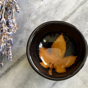 May include: A small, round, black bowl with a preserved orange leaf inside. The bowl is set on a marble surface, with a bundle of dried lavender flowers in the background.