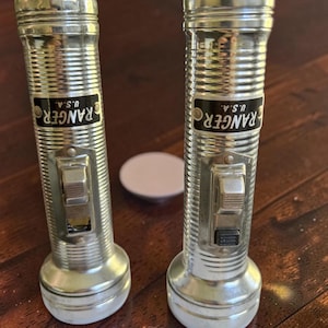 May include: Two vintage silver-toned metal flashlights with ribbed cylindrical bodies. Each flashlight features a black rectangular label with the white text RANGER U.S.A. and a central sliding power switch on the front of the handle.