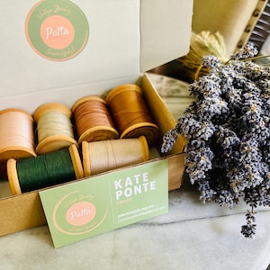 May include: A cardboard box filled with wooden spools of thread in various colors, including beige, green, and brown. A sprig of dried lavender is next to the box. A business card with the text "Kate Ponte" is visible.