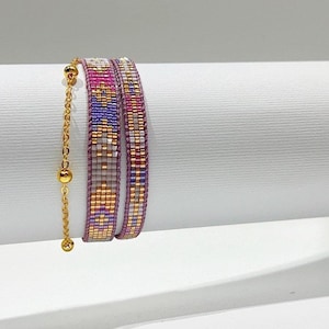 May include: Two beaded bracelets with a gold chain. The bracelets are made of small, colourful beads in shades of pink, purple, and white. The chain has gold beads.