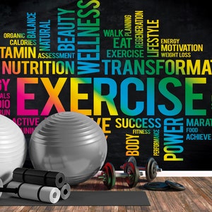 May include: A black chalkboard with colorful text about fitness and exercise. The text includes words like "exercise", "transformation", "wellness", "nutrition", "energy", "motivation", "success", "power", "marathon", "achieve", "focus", "resolution", "sport", "body", "fitness", "goals", "cardio", "run", "active", "walk", "planning", "regeneration", "lifestyle", "eat", "organic", "balance", "natural", "beauty", "calories", "vitamin", "assessment", "fruit", "weight loss". There are two exercise balls, one white and one gray, and a set of dumbbells on a black yoga mat.