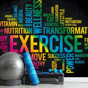 May include: A black chalkboard with colorful text about fitness and wellness. The text includes words like "exercise", "transformation", "nutrition", "energy", "goals", "cardio", "run", "move", "success", "power", "marathon", "achieve", "focus", "resolution", "sport", "fitness", "body", "performance", "food", "organic", "balance", "natural", "beauty", "wellness", "calories", "vitamin", "assessment", "fruit", "planning", "regeneration", "lifestyle", "walk", "eat", "motivation", "weight loss". There is a gray exercise ball, a blue yoga mat, and a black step platform with dumbbells on it in the foreground.