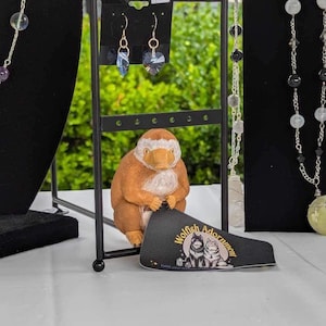 Puede incluir: Un expositor de collares y pendientes hechos con diversas piedras y cristales. Los collares están en soportes negros y los pendientes en un fondo blanco. Las etiquetas de precio son visibles en la imagen. El texto en la pegatina dice "Wolfish Adornments".