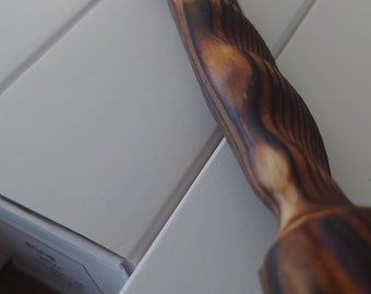 Puede incluir: Una varita de madera con un patrón de veta de madera marrón oscuro y negro. La varita está tallada con detalles intrincados y tiene una punta puntiaguda.