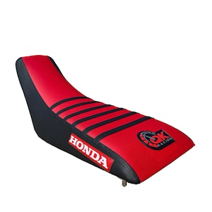 May include: A red and black motorcycle seat with the word "HONDA" in white. The seat has black stripes and a "GK Racing" logo. The seat is designed for a motorcycle.