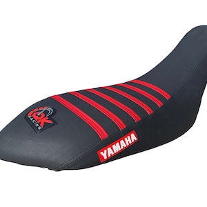 Può includere: Sella da moto nera con strisce rosse e logo "YAMAHA". La sella ha un logo "CK Racing" sul davanti.