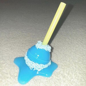 May include: A blue, star-shaped candy with a yellow straw. The candy is decorated with white sprinkles around the base of the straw. The candy is on a light-colored surface.