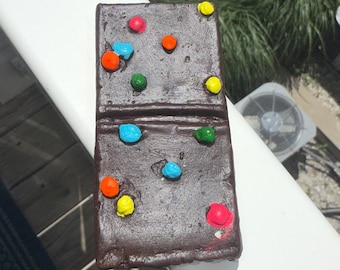 May include: A dark brown domino with colorful, round, raised dots. The dots are in shades of red, orange, yellow, green, and blue. The domino is resting on a white surface.
