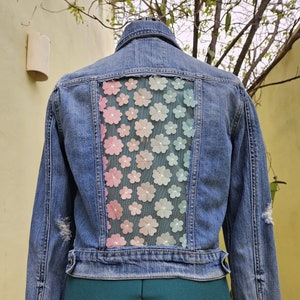 May include: A blue denim jacket with a sheer panel on the back featuring a floral design in shades of pink, blue, and green.