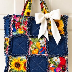 May include: A tote bag constructed from quilted denim squares and floral fabric squares with frayed edges. The bag features a sunflower and bird print, two matching fabric handles, and a large, solid white fabric bow attached to the top right.