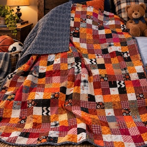 May include: A patchwork quilt featuring a grid of squares in orange, red, black, white, and grey. The fabric squares display various patterns including sports balls, zigzags, and plaid. The reverse side is a solid grey with a repeating pattern.