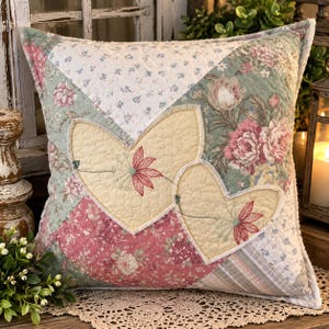 May include: A square quilted throw pillow featuring a patchwork design of floral patterns in shades of pink, green, and white. Two cream-colored heart-shaped fabric appliqués are centered on the front, each embroidered with a red flower.