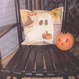 May include: A square decorative cushion featuring a white ghost appliqué with floral patterned eyes and a pumpkin, accented by a cream lace bow, placed next to an orange ceramic jack-o'-lantern on a black wooden slatted rocking chair.