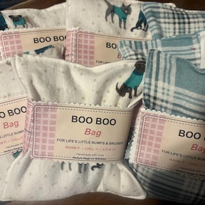 May include: A collection of square fabric boo boo bags featuring white fabric with a pattern of dogs wearing teal sweaters and a blue and white plaid fabric. Each bag has a paper label that reads BOO BOO Bag FOR LIFE'S LITTLE BUMPS & BRUISES.