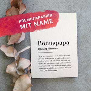 Könnte beinhalten: Eine rechteckige cremefarbene Karte mit dem schwarzen Aufdruck Bonuspapa, Manuel, Substantiv und einer deutschen Definition. Ein roter Pinselstrich mit dem weißen Text PREMIUMPAPIER MIT NAME liegt über dem oberen Bereich. Getrocknete Eukalyptusblätter.