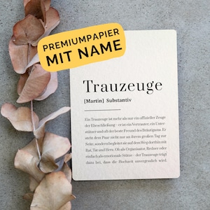 Könnte beinhalten: Eine rechteckige, cremeweiße Karte mit schwarzem Text, der das Wort Trauzeuge definiert, inklusive des Namens Martin. Oben befindet sich ein gelbes Etikett mit dem Text PREMIUMPAPIER MIT NAME. Links liegen getrocknete braune Eukalyptusblätter.