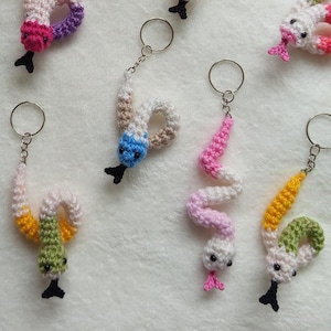 May include: A collection of 18 crocheted snake keyrings in various colours, including pink, blue, green, yellow, orange, and white. Each keyring features a small, crocheted snake with a black bead for an eye.
