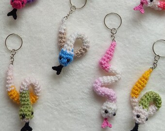 Puede incluir: Una colección de 18 llaveros de serpiente de crochet en varios colores, incluyendo rosa, azul, verde, amarillo, naranja y blanco. Cada llavero presenta una pequeña serpiente de crochet con una cuenta negra para el ojo.