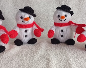 Puede incluir: Cuatro adornos de muñeco de nieve de punto blanco con sombreros negros y bufandas rojas. Los muñecos de nieve están sentados sobre una superficie blanca y esponjosa.