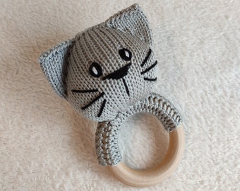 May include: A gray knitted cat rattle with a wooden ring. The cat has black eyes and a black nose and whiskers.