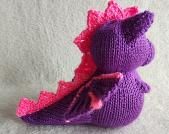 May include: A purple knitted dragon toy with pink crocheted wings and a pink crocheted crown. The dragon is sitting on a white fuzzy surface.