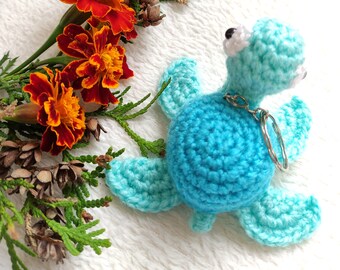 May include: Two crocheted blue sea turtle keychains with white eyes and silver key rings. The turtles are on a white fuzzy surface with a sprig of green foliage and orange marigold flowers.