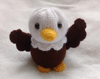 Puede incluir: Un juguete de peluche de águila tejido con plumas blancas, alas marrones y un pico amarillo. El juguete tiene ojos negros y está de pie sobre una superficie blanca.