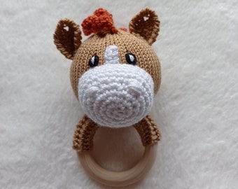 May include: A brown knitted horse head rattle with a white crocheted nose and orange mane. The rattle has a wooden ring attached.