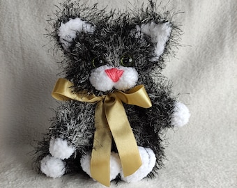 May include: A black and white fluffy crocheted cat toy with a gold ribbon around its neck. The cat has green eyes and a pink nose.