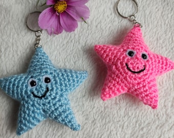 May include: Two crocheted star-shaped keychains, one light blue and one pink, each with a smiling face and a silver keyring. A pink flower with a yellow center is in the background. The keychains are on a white, textured surface.