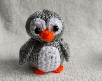 Puede incluir: Un pequeño pingüino de crochet gris y blanco con patas y pico naranja. El pingüino tiene ojos negros y una barriga blanca.