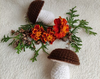 May include: Two crocheted mushroom keychains with brown caps and white stems. The keychains are surrounded by orange marigold flowers and green foliage.