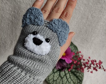 May include: Gray knitted fingerless gloves with a fuzzy gray and blue bear face on the back of the hand.