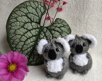 Puede incluir: Dos osos koala de punto gris con pelaje blanco en el vientre y las orejas. Los osos están sentados sobre una superficie blanca y peluda con una hoja verde y flores rosas en el fondo.