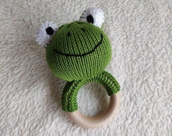 May include: Green knitted frog rattle with a wooden ring. The frog has white eyes and a black smile.