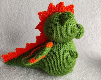 May include: A handmade, knitted green dinosaur toy with orange accents. The dinosaur has a spiky orange crest and tail, with small orange details on its arms. The toy is made of soft yarn and has a friendly, rounded shape.