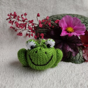 May include: A green knitted frog keyring with white eyes and a black smile. The frog is sitting on a white fluffy surface with pink flowers in the background.