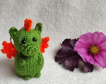 May include: A green crocheted dragon keychain with orange wings and a red horn. The dragon has black eyes and a small, round body.