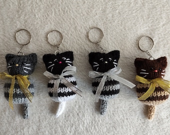 Puede incluir: Cuatro llaveros de gato tejidos con cuerpos rayados y lazos de cinta dorada. Los gatos son grises, blanco y negro, blanco y marrón, y marrón.