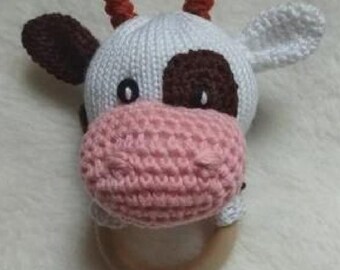May include: A handmade crochet baby rattle shaped like a cow. The cow has a white head with brown spots, brown ears, and orange horns. The muzzle is pink, and the rattle has a wooden ring base.