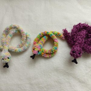 Puede incluir: Tres juguetes de serpiente de crochet. Uno es una serpiente de color rosa claro y blanco, otro es una serpiente de colores arcoíris y el último es una serpiente de color morado oscuro.