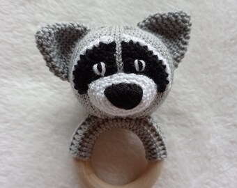 Puede incluir: Un sonajero de mapache de crochet gris con ojos negros y nariz negra. El sonajero tiene un anillo de madera.