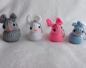 May include: Four knitted bunny keychains in gray, white, pink, and blue. Each bunny has black eyes, an orange nose, and a silver keychain. The bunnies are sitting on a white, textured surface.