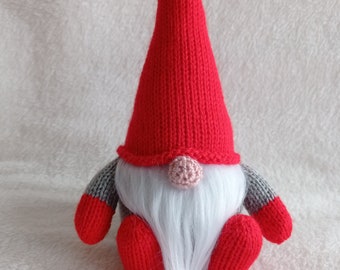 May include: A red knitted gnome with a white beard and a pink nose. The gnome is sitting on a white fluffy surface.
