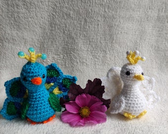 May include: Two crocheted peacocks, one blue with a green tail and one white with a white tail, are sitting on a white fuzzy surface. A pink flower is between the peacocks.