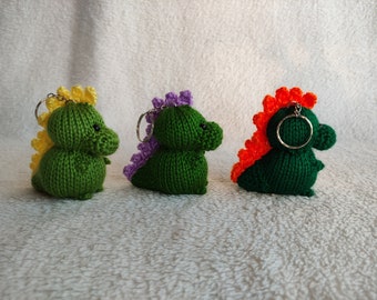 May include: Three knitted green dinosaur keychains with different colored spikes. The dinosaurs have black eyes and are attached to silver key rings.